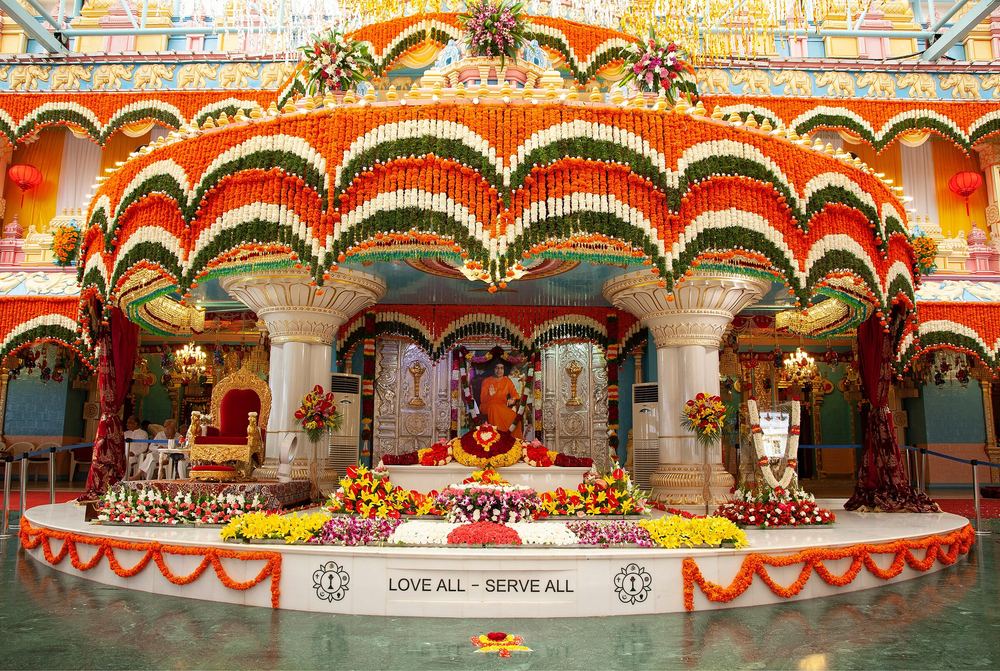 Inside Sai Kulwant Hall — the Mahasamadhi beneath arches of marigold, LOVE ALL — SERVE ALL on the dais (Sri Sathya Sai Media Centre · public domain).