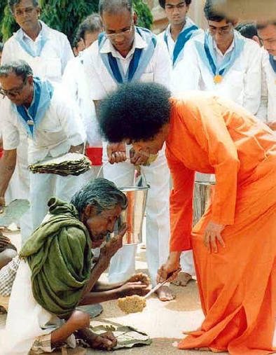 Sri Sathya Sai Baba bends to serve food to a seated elder, Seva Dal volunteers in white and blue behind