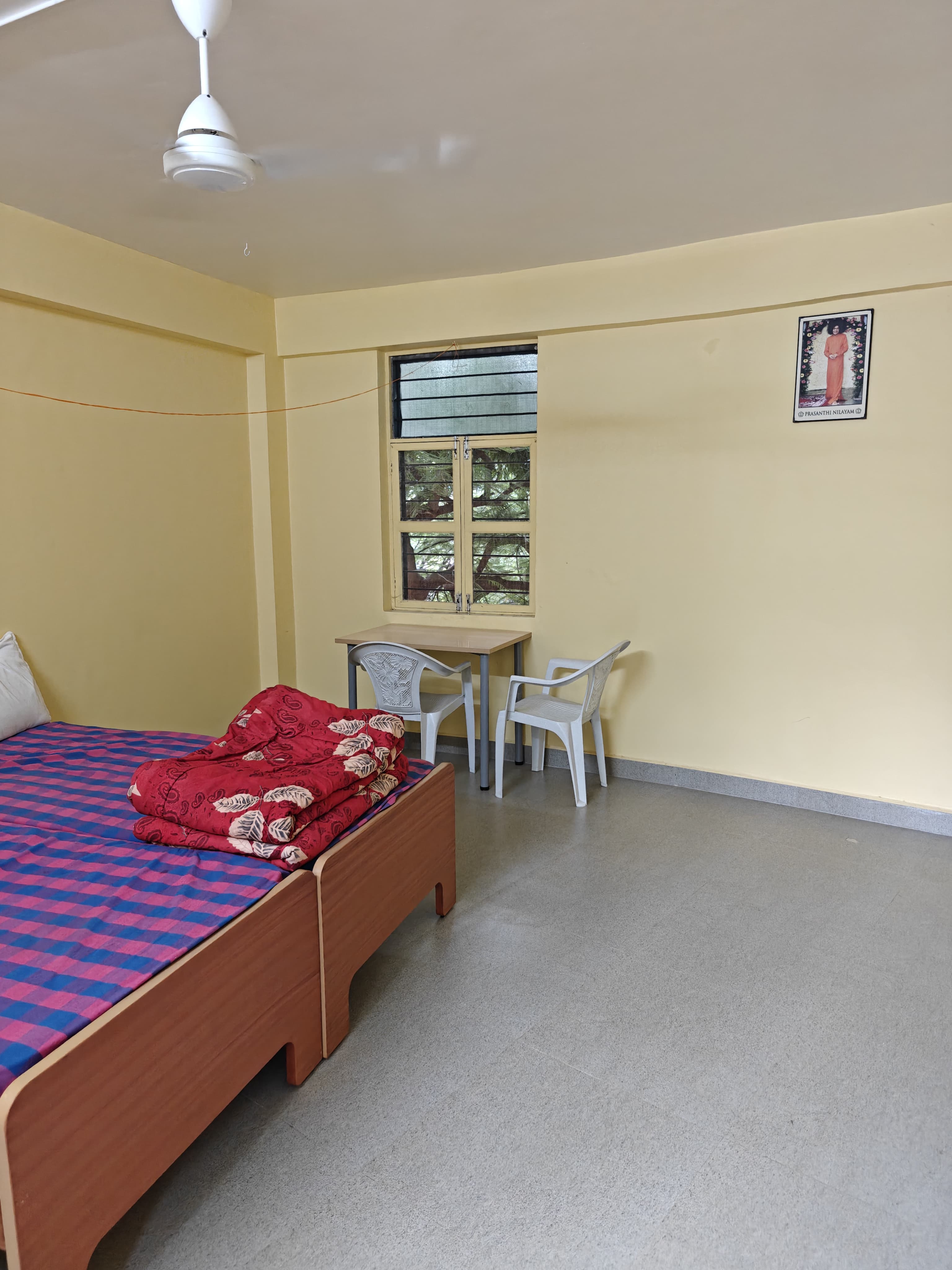 A furnished room at Prasanthi Nilayam: single cot with linen, study table and two chairs, ceiling fan, Baba's photo on the wall