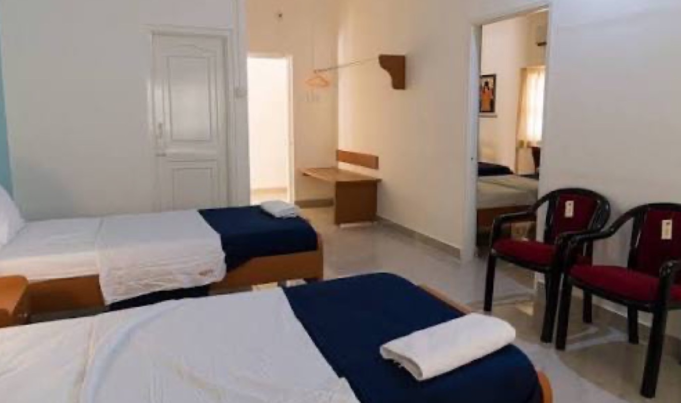 A renovated room with twin cots, seating chairs, a dressing area and attached bathroom